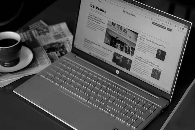 Black and white image of a laptop displaying news articles, accompanied by a cup of coffee and newspapers. HP laptops