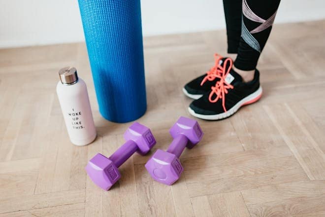 A pair of sneakers, purple dumbbells, yoga mat and bottle on wooden floor. Exercise at Home