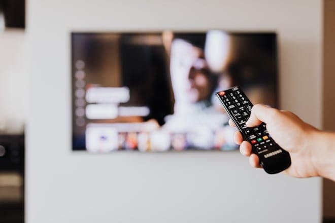 Person using a TV remote control with a blurred television screen in the background. Person using a TV remote control with a blurred television screen in the background.